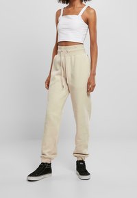 Beige Jogginghose mit elastischen Bündchen und verstellbarem Kordelzugbund, kombiniert mit einem weißen, kurz geschnittenen Tanktop und schwarzen High-Top Sneakers.