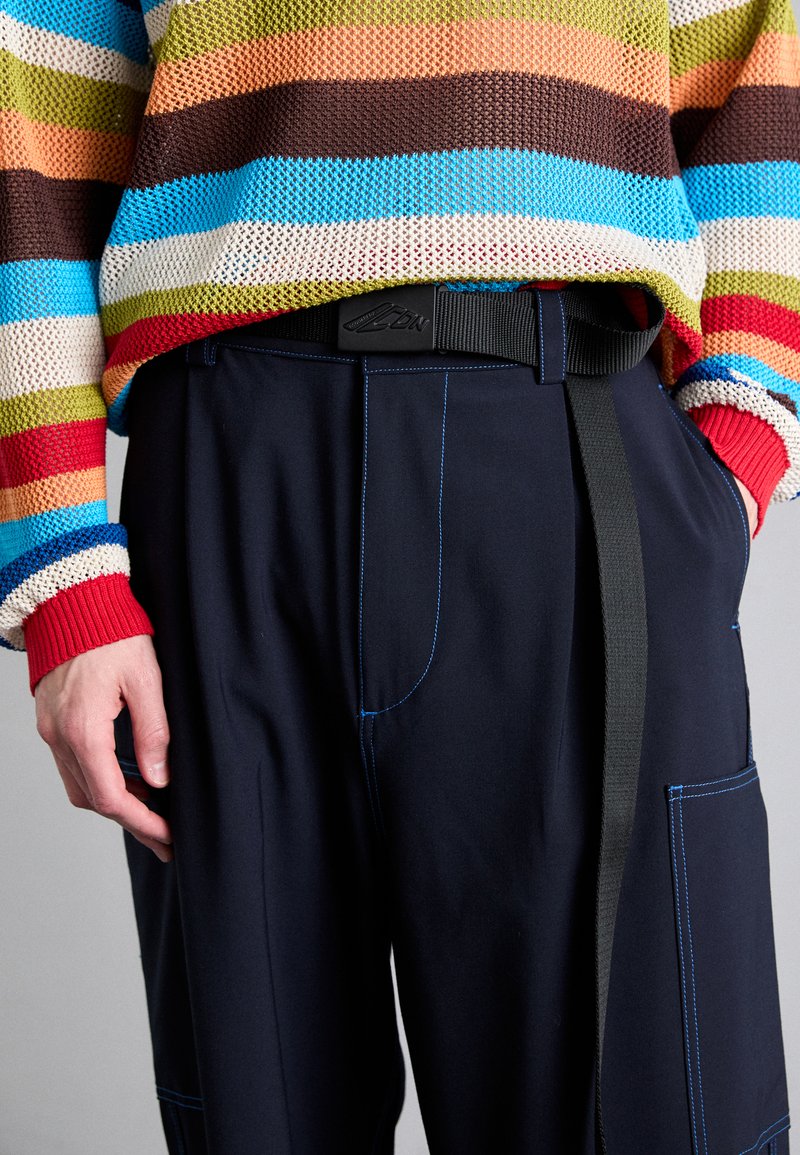 Suéter de punto multicolor con rayas horizontales en azul, verde, rojo y marrón, combinado con pantalones oscuros de color marino con costuras azules y un cinturón.