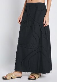 Črna maxi krila ima teksturirano zasnovo z raztrganimi detajli in ukrivljenimi črtami. Nosi se z bež praznimi sandali na eni vezalki.