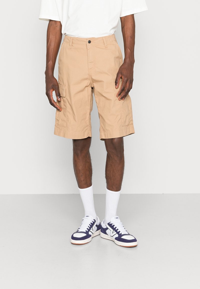 Homme portant un short cargo beige, des chaussettes blanches, des baskets blanches et bleu marine, et une chemise blanche à manches courtes, debout devant un fond uni.