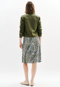 Veste olive à la coupe structurée, associée à une robe midi avec un motif de feuilles vertes et blanches. Chaussures de couleur neutre.