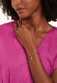 Bracelet en argent avec un band flexible et un petit pendentif "Z", porté sur une main avec un fond de haut rose vif.