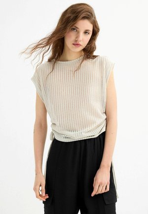 Top sin mangas de crema con textura de canalé vertical y detalle de nudo lateral, combinado con pantalones negros holgados que cuentan con bolsillos.