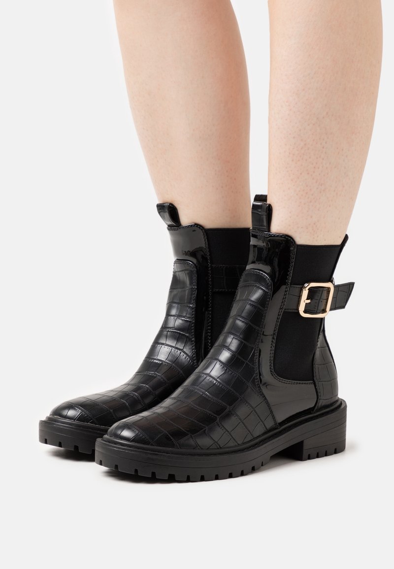 Bottines noires en texture croco avec semelles épaisses, panneaux latéraux élastiques et détail de boucle dorée portées sur jambes nues sur fond blanc.