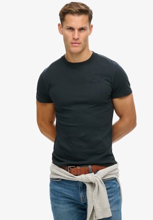 Camiseta negra con cuello redondo, mangas cortas y detalle del logo en púrpura. Jeans de mezclilla azules con un cinturón marrón y un suéter gris atado a la cintura.