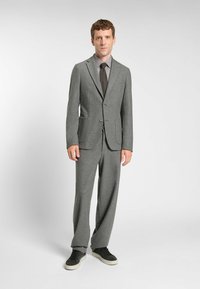 Completo grigio su misura con rever a punta, due tasche frontali, abbinato a una camicia da uomo grigia più chiara e una cravatta marrone. Indossato con sneakers nere casual.