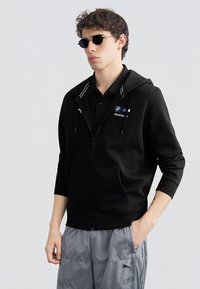 Νεαρός άνδρας που φοράει μαύρο φούτερ BMW Motorsport, μαύρο πόλο, στρογγυλά μαύρα γυαλιά ηλίου και γκρι φόρμα Puma, με το ένα χέρι στην τσέπη.