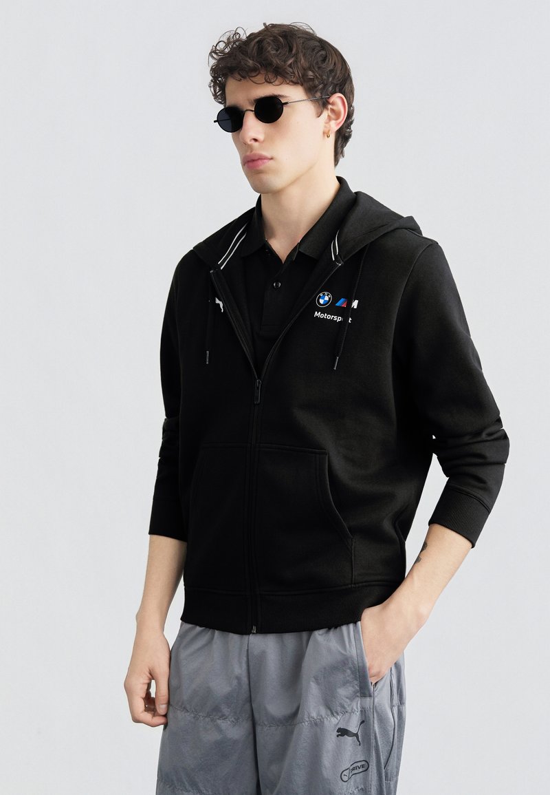 Νεαρός άνδρας που φοράει μαύρο φούτερ BMW Motorsport, μαύρο πόλο, στρογγυλά μαύρα γυαλιά ηλίου και γκρι φόρμα Puma, με το ένα χέρι στην τσέπη.