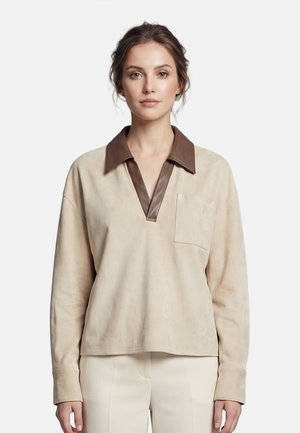 Vrouw met lichtbruin haar naar achter gebonden, draagt een beige top met lange mouwen en een donkerbruine kraag en crèmekleurige broek.