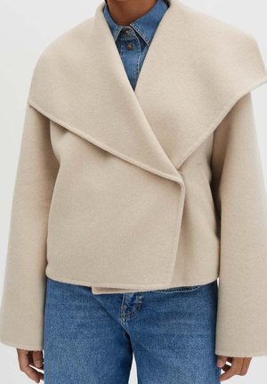 Beige uldfrakke med oversized krave båret over en knappet blå denimskjorte og blå jeans på en person mod en ensfarvet baggrund.