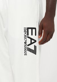 Baltas sporta bikses ar izšūtu melnu logo "EA7 Emporio Armani" un svītru akcentiem kreisajā pusē. Mīksta kokvilnas auduma.