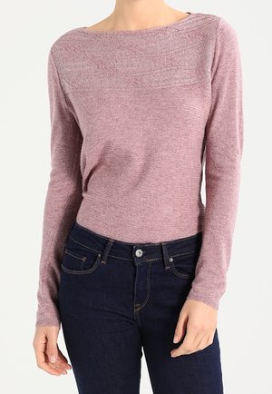 Mujer con un suéter ajustado de manga larga color rosa empolvado con detalles cosidos, combinado con vaqueros azul oscuro sobre un fondo blanco.
