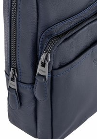 Granatowa skórzana torba z teksturowaną powierzchnią, niebieskimi przeszyciami, dwiema kieszeniami na zamek błyskawiczny oraz metalowymi elementami w kolorze srebrnym do zapięcia.
