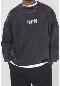 Černá oversized mikina s bílými japonskými znaky na prsou, nošená přes bílou košili, spárovaná s černými kalhotami a stříbrným řetízkem.