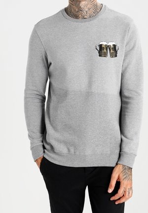 Camiseta gris de manga larga con un gráfico de dos jarras de cerveza brindando en el pecho izquierdo, usada por una persona tatuada con pantalones negros.