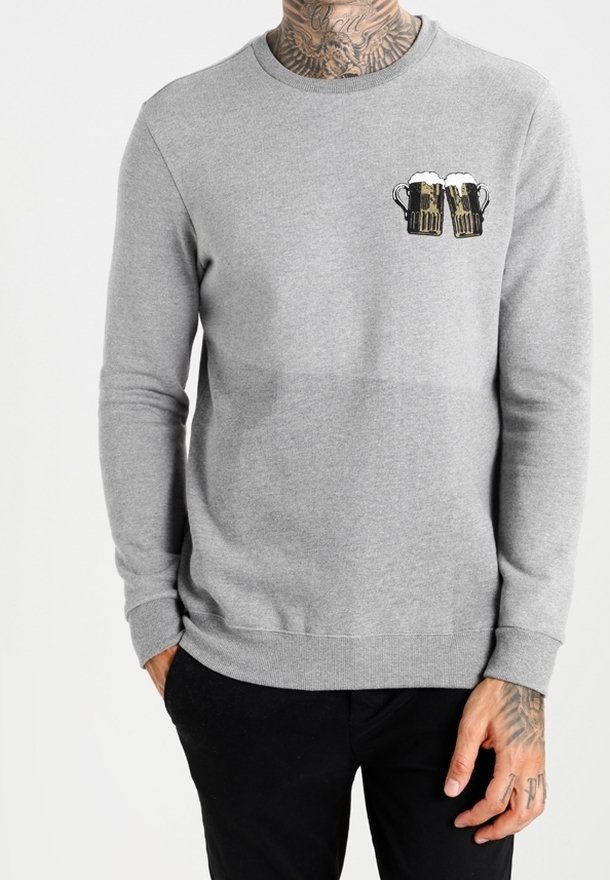 Maglietta grigia a maniche lunghe con grafica di due boccali di birra che si toccano sul petto a sinistra, indossata da una persona tatuata con pantaloni neri.