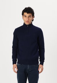 Tamno plavi dukserica na zip s visokim ovratnikom, izrađena od mekanog pletenog materijala. Ima rebraste manžete i donji rub. Diskretni logotip na prsima.