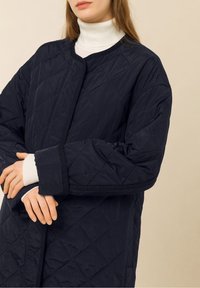 Chaqueta acolchada azul marino con cuello redondo, que presenta una textura suave y puños inclinados. Se lleva sobre una camiseta de cuello alto blanca.