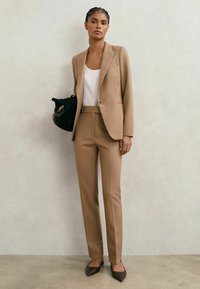 Femeie purtând un costum bej cu pantaloni și sacou asortați, bluză albă, pantofi maro cu vârf ascuțit, ținând o geantă neagră în fața unui perete simplu.