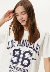T-shirt en coton blanc avec une grande impression bleu marine indiquant "LOS ANGELES 96" accompagnée de "SUPERIOR" et "1999". Coupe décontractée et manches courtes.