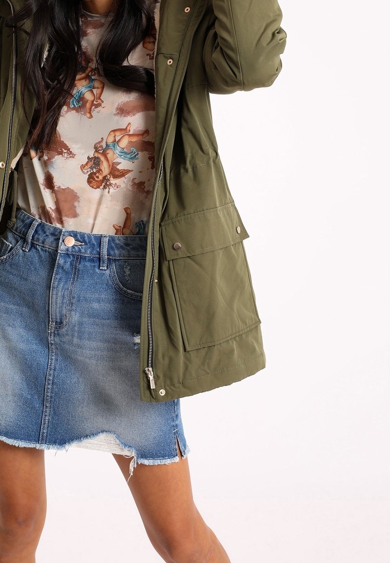 Olijfgroene parka met rits, zijzakken en geribbelde manchetten, gelaagd over een gemusterde blouse en een verouderde denimrok.