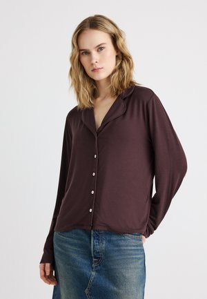 Tânără cu păr blond până la umeri poartă o cămașă maro închis cu mâneci lungi și fustă din denim albastru.