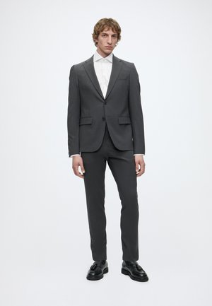 Jeune homme debout portant un costume gris foncé avec une chemise blanche et des chaussures habillées noires sur un fond clair uni.