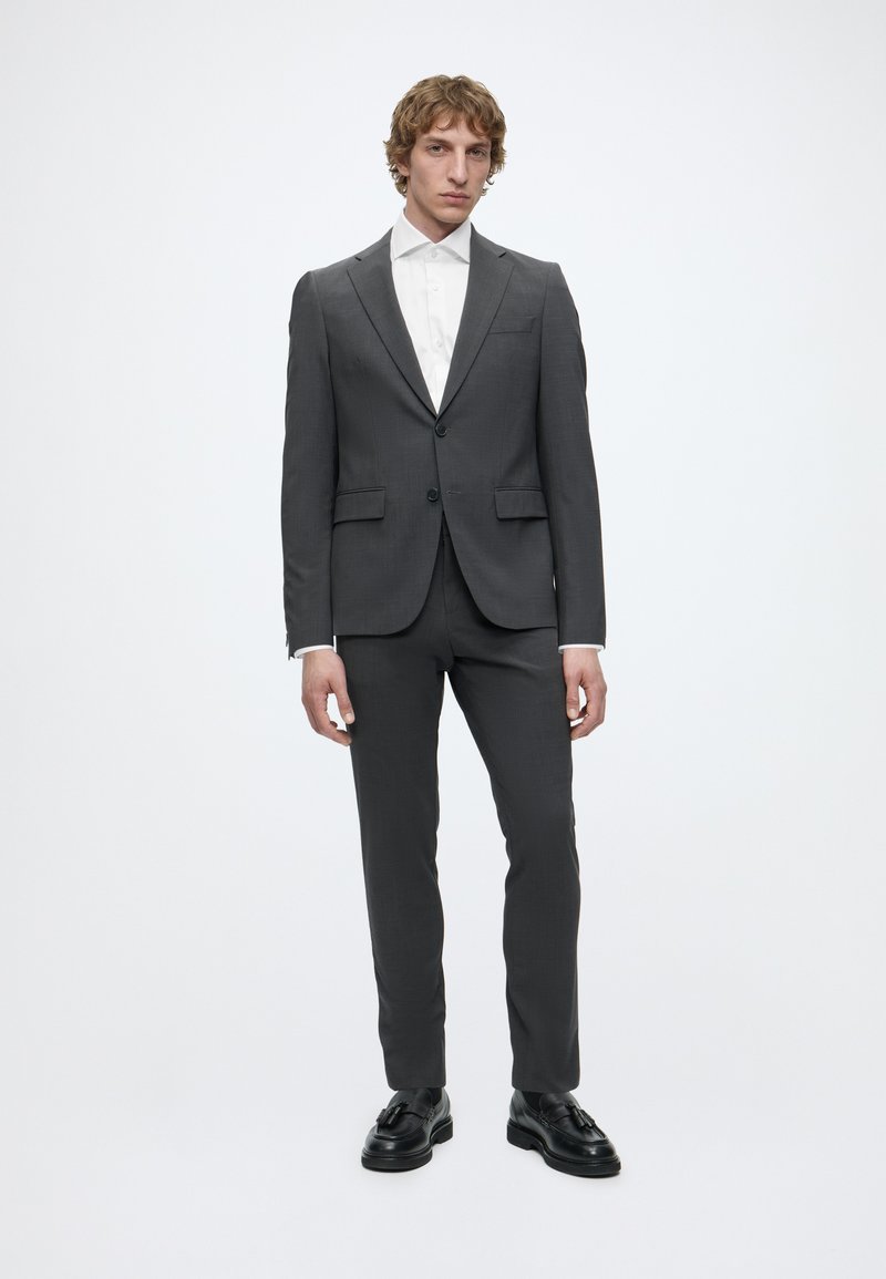 Jeune homme debout portant un costume gris foncé avec une chemise blanche et des chaussures habillées noires sur un fond clair uni.