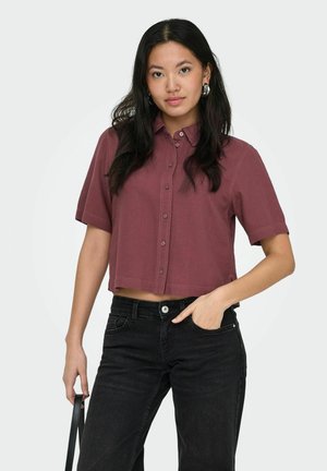 Jeune femme aux longs cheveux foncés portant une chemise boutonnée bordeaux à manches courtes et un jean noir, tenant une lanière de sac à main noire.