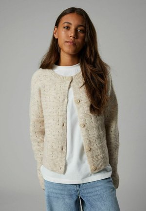 Cardigan beige en tricot avec un motif texturé, doté d'un col rond, de cinq boutons et de poignets côtelés ; porté sur une chemise blanche.