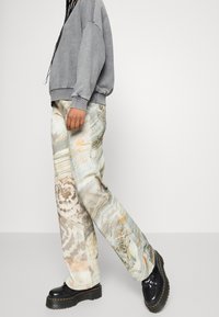 Jasne spodnie z abstrakcyjnym wzorem w odcieniach szarości, brązu i pomarańczu, w połączeniu z luźną szarą bluzą i czarnymi masywnymi butami.