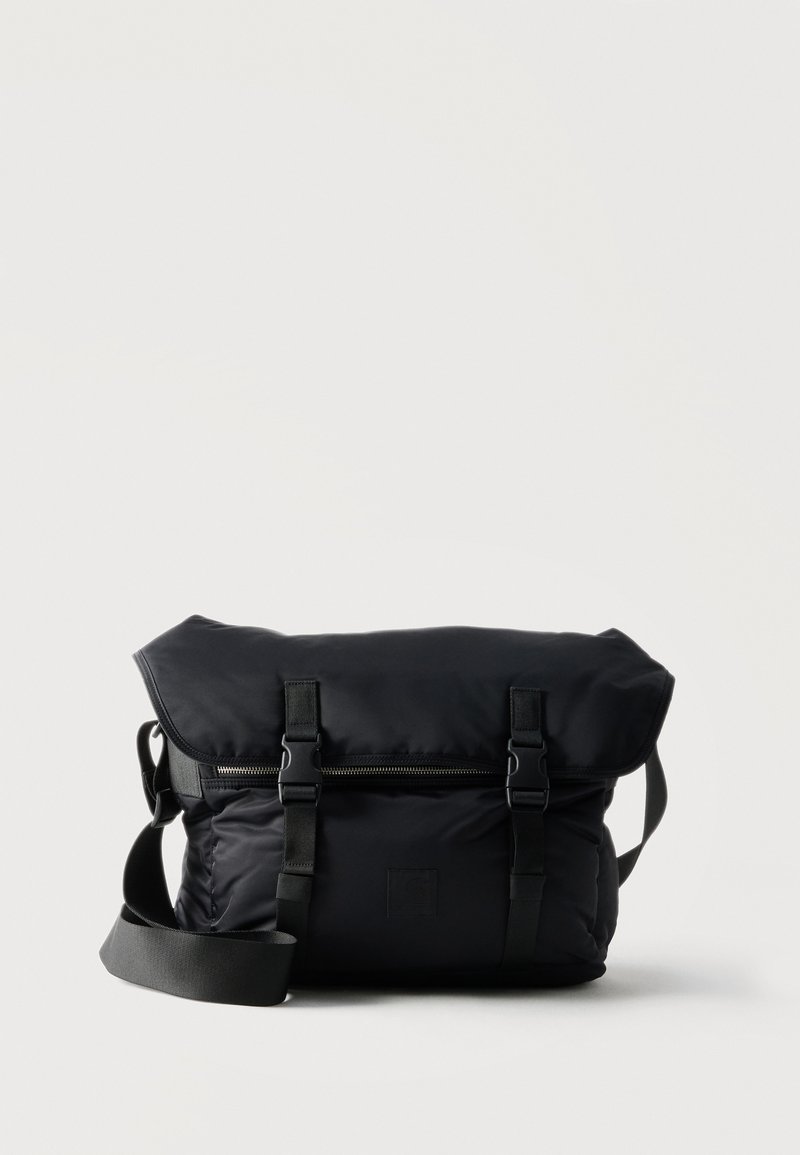 Sac messager en tissu noir avec sangle d'épaule réglable, deux fermetures à boucle à l'avant et une poche zippée sous le rabat.