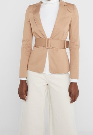 Personne portant un blazer beige ceinturé, un col roulé blanc et un pantalon large crème sur fond uni.