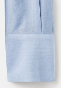 Polsino di camicia color azzurro chiaro con un tessuto testurizzato, orlo pulito e pieghe sottili, che enfatizzano un aspetto liscio e fresco.