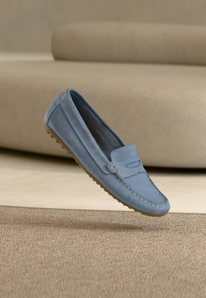 Mocasín de ante azul claro con detalles cosidos y suela de goma, suspendido en el aire frente a un fondo de sofá y alfombra beige.