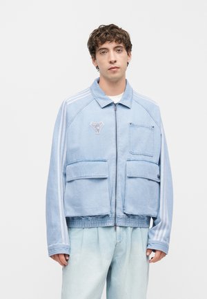 Jeune homme portant une veste zippée bleu clair avec plusieurs poches et des bandes blanches sur les manches, associée à un pantalon bleu clair.