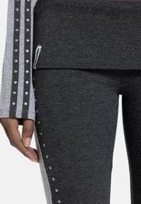 Κοντινό πλάνο χεριού που ακουμπά δίπλα σε σκούρο γκρι παντελόνι με ασημί τρουκς κατά μήκος της πλαϊνής ραφής και γκρι μανίκι με ριγέ τρουκς.