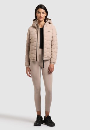 Kobieta w beżowej pikowanej kurtce z kapturem, beżowych legginsach, czarnej bluzce i czarnych trampkach, stojąca na jasnoszarym tle.