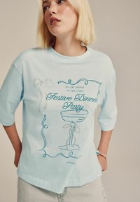 Junge Frau mit kurzem blonden Haar, die ein hellblaues T-Shirt trägt, auf dem ein festlicher Partyeinladungstext und eine Linzeichnung eines Cocktailglases zu sehen sind.