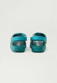 Tealfärgade open-toe sandaler med justerbara remmar och ett vitt tryckt mönster på sulan, visade bakifrån på en enkel bakgrund.