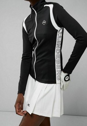 Schwarz schmal geschnittene Jacke mit weißen Seitenstreifen und dem Schriftzug "J.LINDEBERG", kombiniert mit einem weißen plissierten Rock und einem Golfhandschuh an einer Hand.