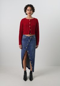 Cardigan court en tricot rouge avec col rond, associé à une jupe en denim taille haute avec une fente à l'avant et des boutons décoratifs. Bottines noires.