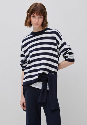 Junge Frau mit kurzen braunen Haaren, die einen marineblauen und weißen, horizontal gestreiften Pullover und marineblaue Hosen trägt, posiert vor einem schlichten grauen Hintergrund.