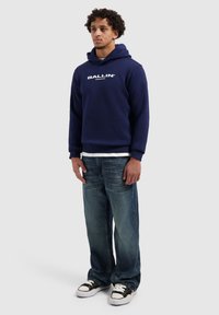 Marineblauwe hoodie met de tekst "BALLIN", gecombineerd met lichtblauwe spijkerbroek en zwarte sneakers. Casual ontwerp, comfortabele pasvorm, zachte stof.