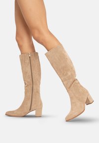 Botas de gamuza beige hasta la rodilla con tacón bloque y cremallera lateral. El diseño tiene una textura suave y punta afilada.