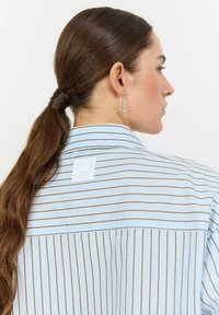 Ljusblå randig skjorta med bruna vertikala linjer och en liten logotags på baksidan. Slät tyg och kragdesign.