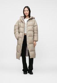 Beige, knee-length puffer coat with a hood, featuring a zip front, large quilted sections, and drawstrings. Worn over black pants and shoes.