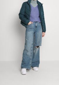 En blågrøn puffjakke, lysegrå rullekrave, lilla strikket vest, vidde bukser med slid og hvide sneakers udgør et afslappet lag-på-lag outfit.