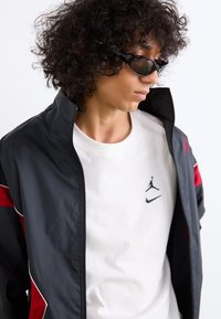 Veste noire et rouge avec fermeture éclair et col montant, accompagnée d'un t-shirt blanc arborant les logos noirs de Nike et Jordan.