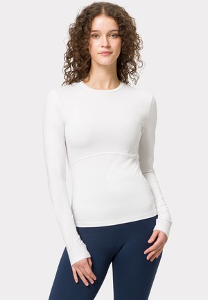 Haut de sport blanc à manches longues, au design ajusté, texture lisse, avec un détail de couture incurvée à la taille, associé à un legging sombre.